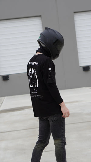 Person wearing a black helmet and black shirt with white graphics and text, standing in front of a gray warehouse.