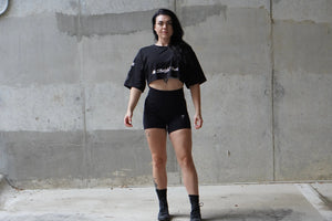 Woman wearing a black crop top and shorts standing against a concrete wall.