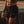 A shirtless, muscular man wearing black athletic shorts and a black wristwatch, standing on an outdoor track or field with a dark tarp in the background.