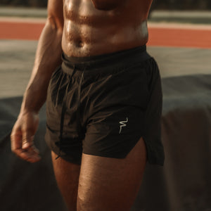 A shirtless, muscular man wearing black athletic shorts and a black wristwatch, standing on an outdoor track or field with a dark tarp in the background.