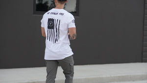 Person wearing a white t-shirt with a design on the back, standing against a dark wall.