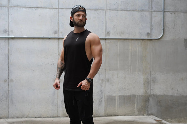 Man wearing a black tank top and cap standing against a concrete wall.