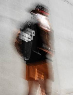 A person wearing a black shirt with a large white 'NSP' logo on the back, paired with orange shorts and a dark cap, captured in a motion-blurry double-exposure style against a light gray concrete wall.