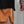 Close-up of a person wearing orange athletic shorts with white vertical 'No STRAIGHT PATH' branding, paired with a black top against a gray concrete wall.