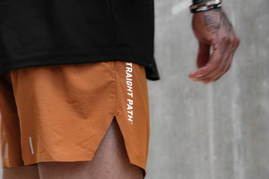 Close-up of a person wearing orange athletic shorts with white vertical 'No STRAIGHT PATH' branding, paired with a black top against a gray concrete wall.