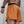 A close-up back view of a man wearing orange athletic shorts with a black waistband, standing against a textured gray concrete wall.