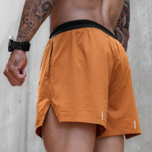 A close-up back view of a man wearing orange athletic shorts with a black waistband, standing against a textured gray concrete wall.