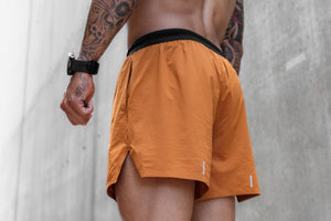 A close-up back view of a man wearing orange athletic shorts with a black waistband, standing against a textured gray concrete wall.