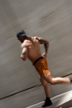 A shirtless man running in orange athletic shorts and a black cap, captured with motion blur against a gray concrete background.