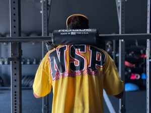 Person wearing a yellow 'NSP' shirt lifting weights in a gym.