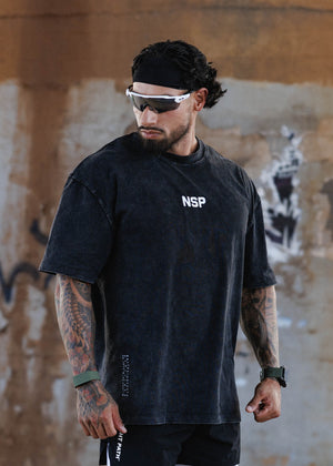 A man wearing a black mineral-washed t-shirt with a white 'NSP' logo on the chest, black athletic shorts, and a black headband, standing against a textured concrete wall.