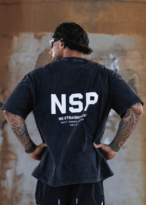 A man viewed from behind wearing a faded black t-shirt with 'NSP' printed in white on the back, paired with black athletic shorts over white leggings, standing against a textured concrete wall.