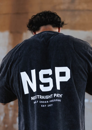 A person viewed from behind wearing a dark washed black t-shirt with a large white 'NSP' logo and 'NOSTRAIGHT PATH BUILT UNDER PRESSURE EST 2021' printed on the back, set against a rustic wooden background.