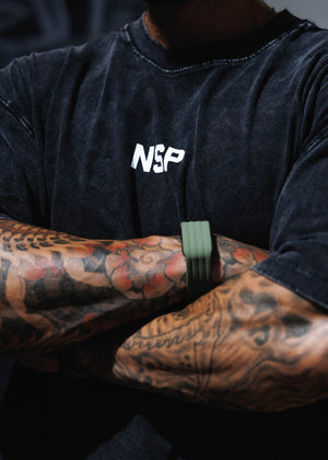 Close-up of a tattooed person wearing a faded black t-shirt with a white 'NSP' logo on the chest, paired with an olive green silicone wristband.