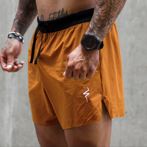 A shirtless tattooed man wearing orange athletic shorts with a black waistband, a black cap, and red-tinted sunglasses, standing against a concrete wall.