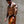 A shirtless tattooed man wearing orange athletic shorts, a black cap, and red-tinted sunglasses walks against a concrete wall.