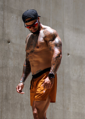 A shirtless tattooed man wearing orange athletic shorts, a black cap, and red-tinted sunglasses walks against a concrete wall.
