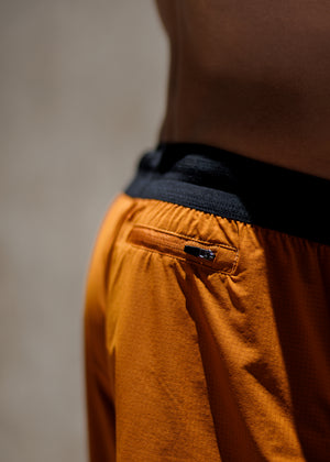 Close-up of a person wearing orange athletic shorts with a black waistband and a small black zipper pocket detail, photographed against a neutral beige background.